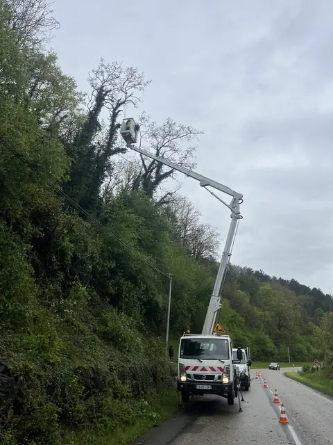 En nacelle au-dessus du talus boisé, la route balisée par des cônes.